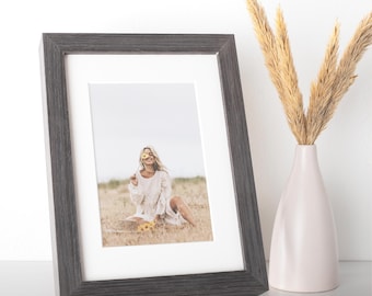 May include: A rectangular picture frame with a dark grey wood grain finish and a white mat board. The frame contains a photograph of a person in a white dress sitting in a field, holding a yellow flower. Next to the frame is a light pink vase.