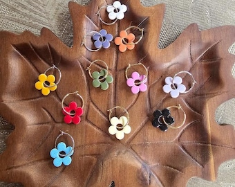 May include: A collection of flower-shaped hoop earrings in various colors, including white, purple, orange, yellow, green, pink, red, light blue, cream, and black. The earrings are displayed on a wooden leaf-shaped tray.