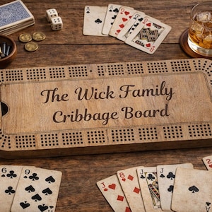 Peut inclure: Un jeu de société de cribbage en bois avec l'inscription "The Wick Family Cribbage Board" est entouré de cartes à jouer, de dés et de pièces de monnaie. Un verre de whisky est posé sur un dessous de verre.