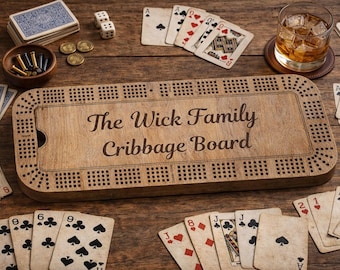 May include: A wooden cribbage board with the text "The Wick Family Cribbage Board" is surrounded by playing cards, dice, and coins. A glass of whiskey sits on a coaster. The board is a light brown color.