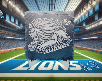 May include: A slate coaster with a white Detroit Lions logo featuring a lion with a mane and the words "Detroit Lions". The coaster is sitting on a wooden stand in front of a football stadium.
