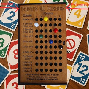 Könnte beinhalten: Holzbrettspiel mit dem Text "PAPA'S 10 Phases" und bunten Spielsteinen. Das Brett hat Löcher für die Spielsteine und ist von Spielkarten mit Zahlen und Farben umgeben. Das Spielbrett hat eine hellbraune Farbe.