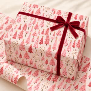 May include: A rectangular gift box wrapped in light pink paper featuring a repeating pattern of watercolor-style pink and red Christmas trees with gold stars, tied with a dark red velvet ribbon finished in a bow on the top right corner.
