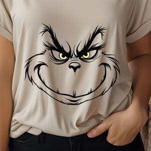 Pode incluir: Uma camiseta bege com uma ilustra&ccedil;&atilde;o em preto e branco do rosto do Grinch. O Grinch tem um sorriso travesso e olhos amarelos.