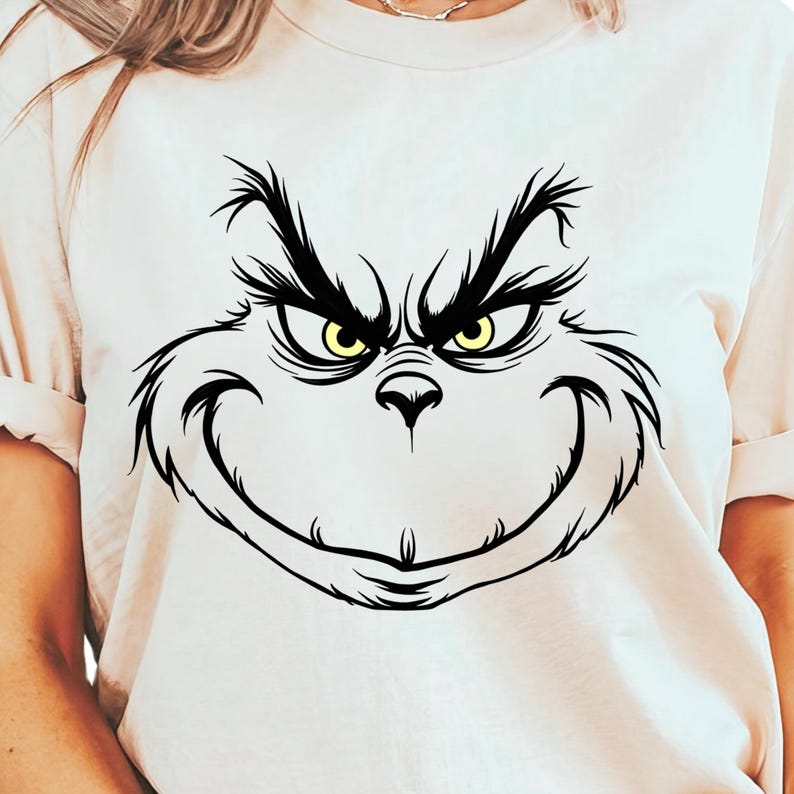 Pode incluir: Uma camiseta branca com uma ilustra&ccedil;&atilde;o em preto e branco do rosto do Grinch. O Grinch tem olhos amarelos e um sorriso travesso.