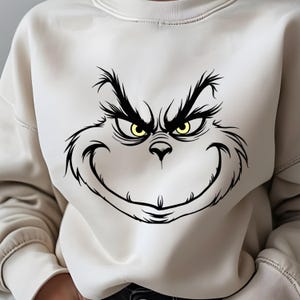 Pode incluir: Uma camisa de moletom branca com uma ilustra&ccedil;&atilde;o em preto e branco do rosto do Grinch. O Grinch tem um sorriso travesso e olhos amarelos.