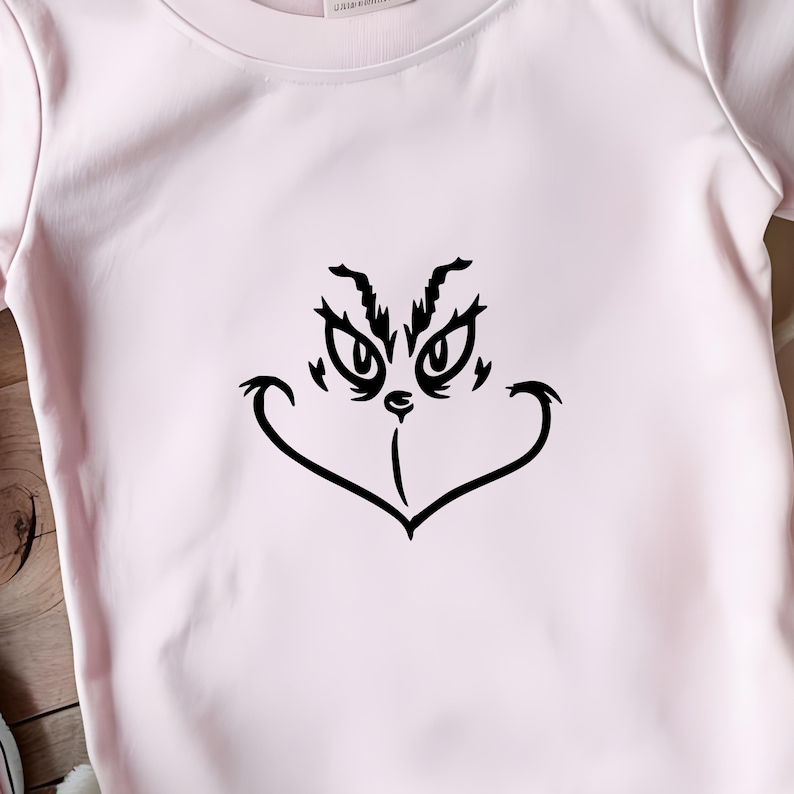May include: A pink t-shirt with a black Grinch face graphic. The Grinch is smiling with his eyes wide open.