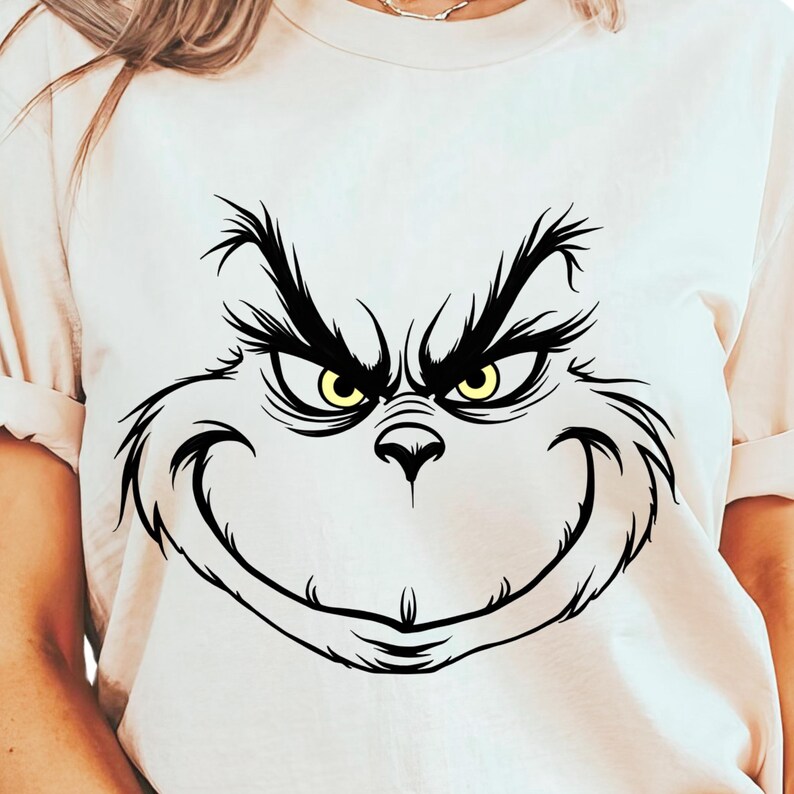 Pode incluir: Uma camiseta branca com uma ilustra&ccedil;&atilde;o em preto e branco do rosto do Grinch. Os olhos do Grinch s&atilde;o amarelos.