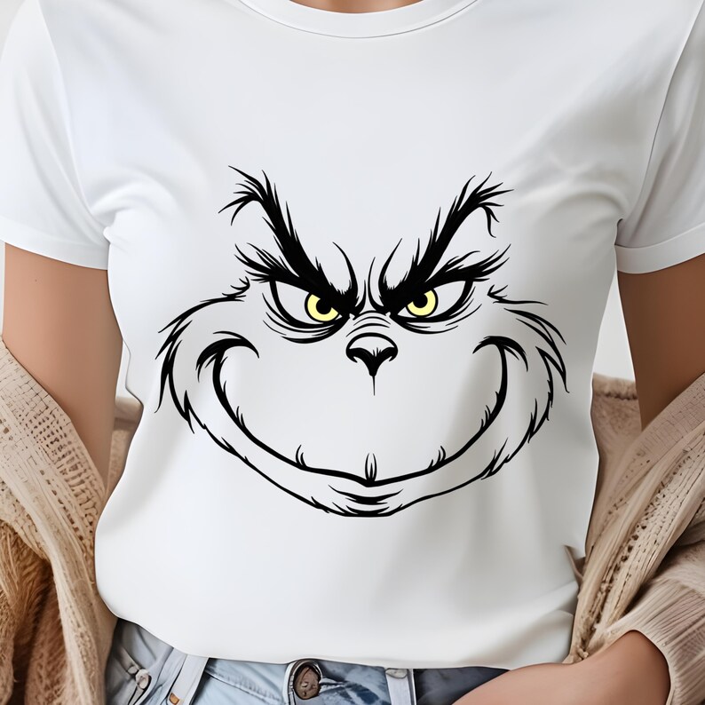 Pode incluir: Camiseta branca com uma ilustra&ccedil;&atilde;o em preto e branco do rosto do Grinch. O Grinch tem um sorriso travesso e olhos amarelos.