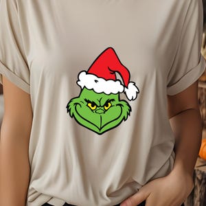 May include: A beige t-shirt with a green Grinch wearing a red Santa hat. The Grinch is looking at the viewer with a mischievous expression.