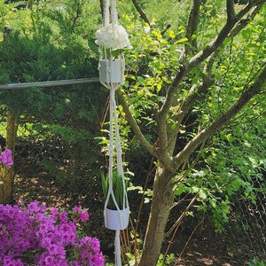 May include: A white macrame plant hanger with two white ceramic pots, one holding white flowers and the other with green grass. The plant hanger is suspended from a tree branch in a garden setting.