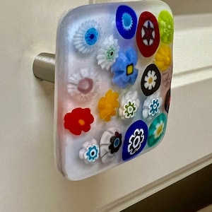 May include: A square cabinet knob with a clear, glass-like body embedded with colorful, floral millefiori designs. The flowers are red, blue, yellow, white, and green. The knob has a brushed metal stem.