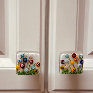May include: Two square cabinet knobs made of clear glass with embedded millefiori floral patterns. Each knob features a white base with colorful glass flowers in shades of red, blue, yellow, and pink, with green stems and foliage at the bottom.