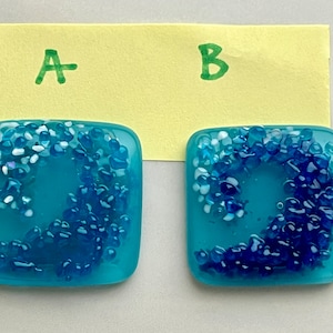 May include: Two square glass art pieces with a wave design. The base color is teal, with a wave pattern made of blue and white glass pieces. The background is a light yellow paper with the letters "A" and "B" written on it.