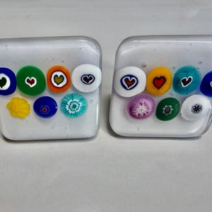 May include: Two square, clear glass decorative objects. Each has a collection of colorful, round, patterned glass pieces embedded within. The patterns include hearts, flowers, and other designs in various colors like blue, green, yellow, and white.