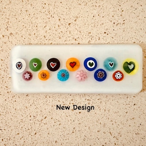 May include: A white rectangular tray displays a collection of colorful, round glass beads. Each bead features a unique design, including hearts, flowers, and geometric patterns. The text "New Design" is visible below the tray.