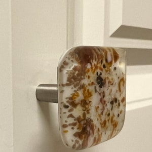 May include: A square cabinet knob with a stainless steel stem. The knob is clear with a speckled design of brown, orange, and black. The knob is mounted on a white cabinet door.