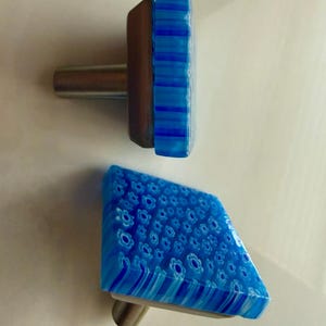 May include: Two decorative cabinet knobs. One knob features a rectangular shape with blue and white striped glass. The other knob has a blue floral pattern. Both knobs have a silver-colored metal stem and a brown metal base.