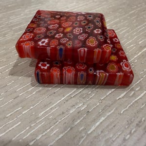 May include: Two square, red glass coasters with a floral millefiori design. The coasters are stacked on top of each other, showcasing the intricate patterns of small, colorful flowers embedded in the glass. The coasters are on a light gray surface.