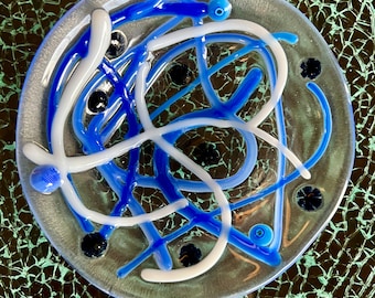 Puede incluir: Un plato de vidrio transparente con un diseño abstracto. El diseño presenta líneas de vidrio azules y blancas entrelazadas, junto con pequeños acentos de vidrio negro. El plato tiene un borde plateado y se encuentra sobre una superficie texturizada de color verde oscuro.