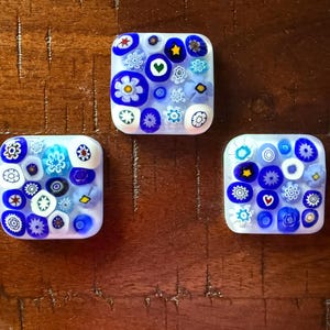 May include: Three square glass tiles with a white base and a colorful floral design. The designs include blue, white, and yellow flowers, stars, and hearts. The tiles are arranged on a dark brown wooden surface.