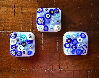 May include: Three square glass tiles with a white base and a colorful floral design. The designs include blue, white, and yellow flowers, stars, and hearts. The tiles are arranged on a dark brown wooden surface.