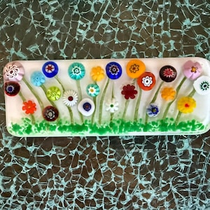 May include: A rectangular fused glass piece featuring a white background with a green grassy base. The design includes numerous colorful millefiori flowers in shades of red, blue, yellow, orange, purple, and white, each with thin green stems.