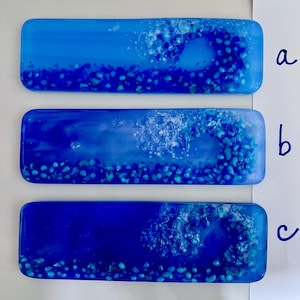 May include: Three rectangular glass art pieces in shades of blue. Each features a wave-like design with small, blue, pebble-like elements. The top piece is light blue, the middle is a mix of blue and white, and the bottom is dark blue.