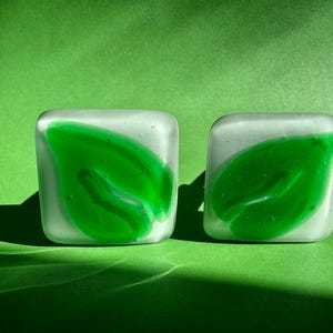 May include: Two square glass tiles with rounded corners, featuring a translucent white base and a vibrant green leaf-shaped design fused into the center. The glass has a smooth, matte finish, creating a soft appearance with distinct green leaf motifs.