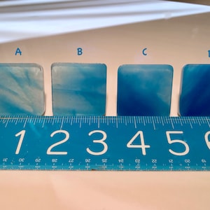 May include: Four square, blue resin pieces with varying shades of blue, arranged in a row. A blue ruler with white markings and numbers from 1 to 6 is placed below the pieces. The pieces are labeled A, B, C, and D.