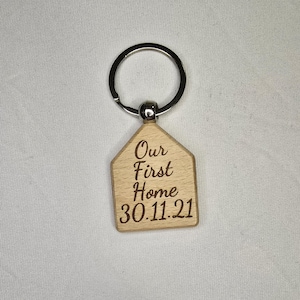 May include: Wooden house-shaped keyring with a silver key ring. The keyring is engraved with the text "Our First Home 30.11.21".