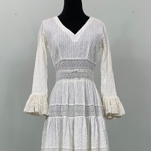 Könnte beinhalten: Ein weißes Maxikleid mit V-Ausschnitt und langen Ärmeln. Das Kleid hat einen Stufenrock mit Spitzenbesatz und ein tailliertes Oberteil. Das Kleid ist aus einem leichten Stoff gefertigt und hat eine lockere Passform.