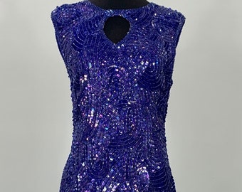 May include: A sleeveless, sequined dress in a deep blue color with a heart-shaped cutout at the neckline. The sequins are iridescent and create a shimmering effect.