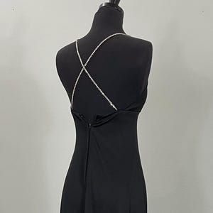 May include: A black evening gown with a sleek silhouette. The dress features a criss-cross rhinestone strap design across the back. The gown is displayed on a black mannequin against a neutral background.