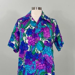 May include: A short-sleeved shirt with a bold floral print. The shirt has a white base, adorned with large flowers in purple, blue, and teal, with green foliage. The garment is displayed on a mannequin, showcasing its design.