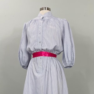May include: A vintage-style dress with vertical blue and white stripes. The dress features a high collar, long sleeves, and a bright pink sash at the waist. The skirt is a midi length. The dress is displayed on a mannequin.