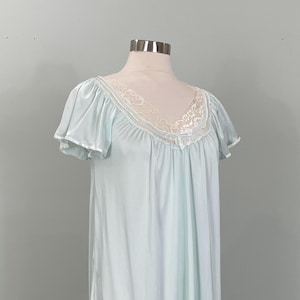 May include: A light blue nightgown featuring short flutter sleeves and a V-neckline trimmed with white floral lace. The garment has a gathered bodice and a loose, flowing silhouette that extends to a mid-length hem, displayed on a mannequin.