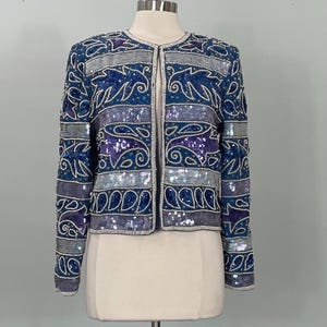 May include: A blue sequined jacket with a round neckline and long sleeves. The jacket features a pattern of swirling designs in shades of blue, purple, and silver. The jacket is displayed on a mannequin.
