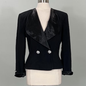Könnte beinhalten: Ein schwarzer Blazer mit Satinrevers und Manschetten. Die doppelreihige Jacke hat zwei dekorative Strassknöpfe und Strassakzente an den Manschetten. Die Jacke wird auf einer Schaufensterpuppe präsentiert.