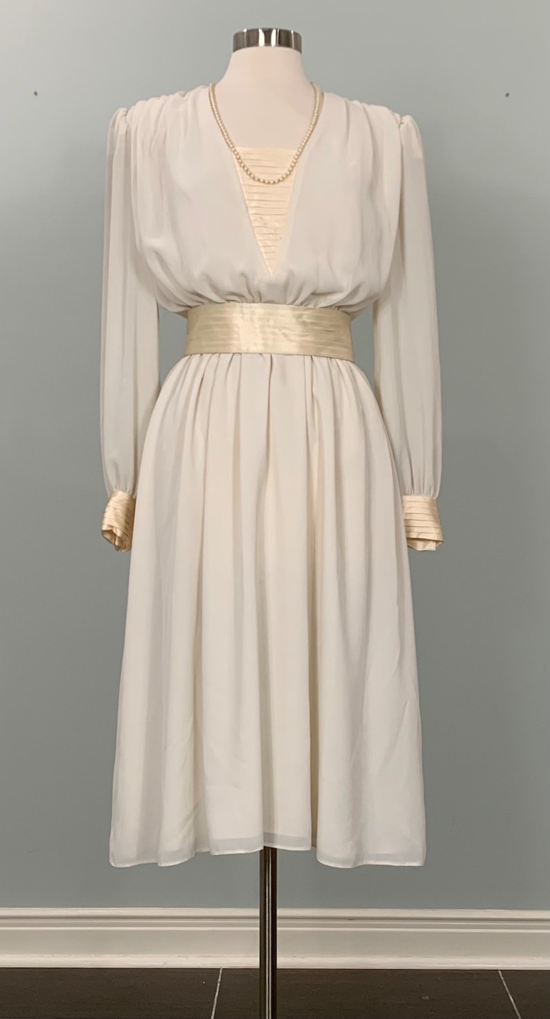 May include: A white vintage dress with a V-neckline, long sleeves, and a satin sash. The dress has a gathered skirt and a pearl necklace.