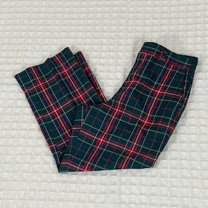 May include: Green plaid pants with red, yellow, and blue accents. The pants are folded and displayed on a white textured surface. The plaid pattern is a classic design.