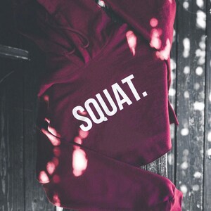 May include: A maroon hoodie with the word "SQUAT" printed in large white letters. The hoodie is made of a soft material and has a drawstring hood. The background is a dark, wooden surface.