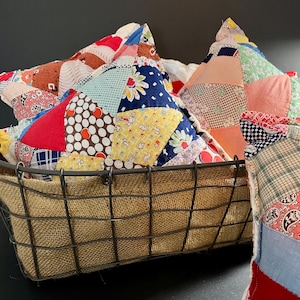 May include: A metal wire basket lined with burlap holds several colorful, patchwork pillows. The pillows feature a variety of patterned fabrics in red, blue, yellow, and white. The basket sits against a dark background.
