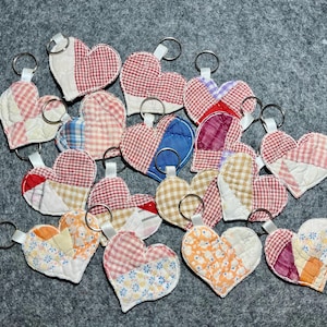 May include: A collection of 16 handmade patchwork heart-shaped keychains. Each keychain is made from different fabrics in shades of red, pink, white, blue, and brown. The keychains are arranged in a semi-circle on a gray surface.