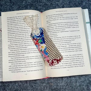 May include: A close-up of a book with a fabric bookmark. The bookmark is made of patchwork fabric with floral patterns and a checkered pattern. The bookmark is tucked into the pages of the book, which is open to a page with text.