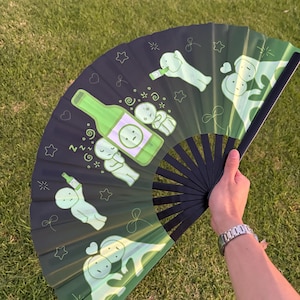 Puede incluir: Un abanico plegable con fondo degradado en negro y verde que presenta personajes de dibujos animados y una botella verde. El dise&ntilde;o incluye varias figuras peque&ntilde;as en diversas posturas, algunas sosteniendo botellas, junto a estrellas.