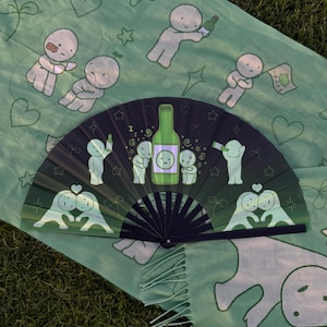 Puede incluir: Un abanico plegable con fondo degradado de verde oscuro a negro, con personajes de dibujos animados, estrellas y una ilustraci&oacute;n central de una botella verde. El abanico se muestra sobre una tela verde claro con un patr&oacute;n de personajes.