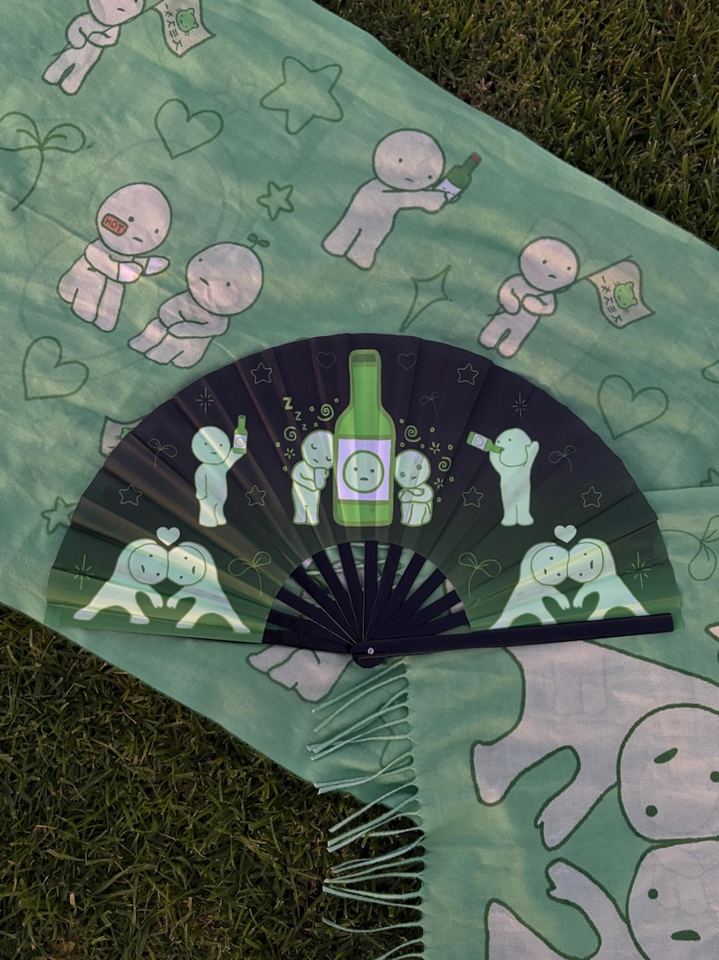 Puede incluir: Un abanico plegable con fondo degradado de verde oscuro a negro, con personajes de dibujos animados, estrellas y una ilustraci&oacute;n central de una botella verde. El abanico se muestra sobre una tela verde claro con un patr&oacute;n de personajes.