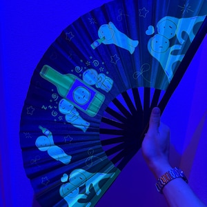 Puede incluir: Un abanico plegable negro con un estampado gr&aacute;fico azul brillante de figuras de dibujos animados y una botella. El dise&ntilde;o incluye varios personajes en diversas posturas, algunos sosteniendo botellas, rodeados de estrellas y l&iacute;neas.