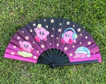 Puede incluir: Un abanico plegable con marco negro y superficie de tela con degradado de morado oscuro a rosa. El abanico presenta varias ilustraciones del personaje rosa Kirby en diversas poses, incluyendo con auriculares y gorro de dormir, rodeado de estrellas.